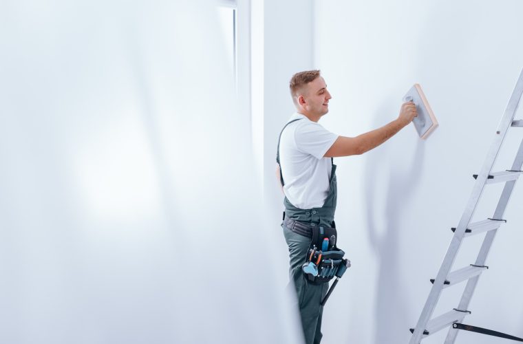 Young smiling painter with tool belt pinned to his workwear smoothing the wall before painting